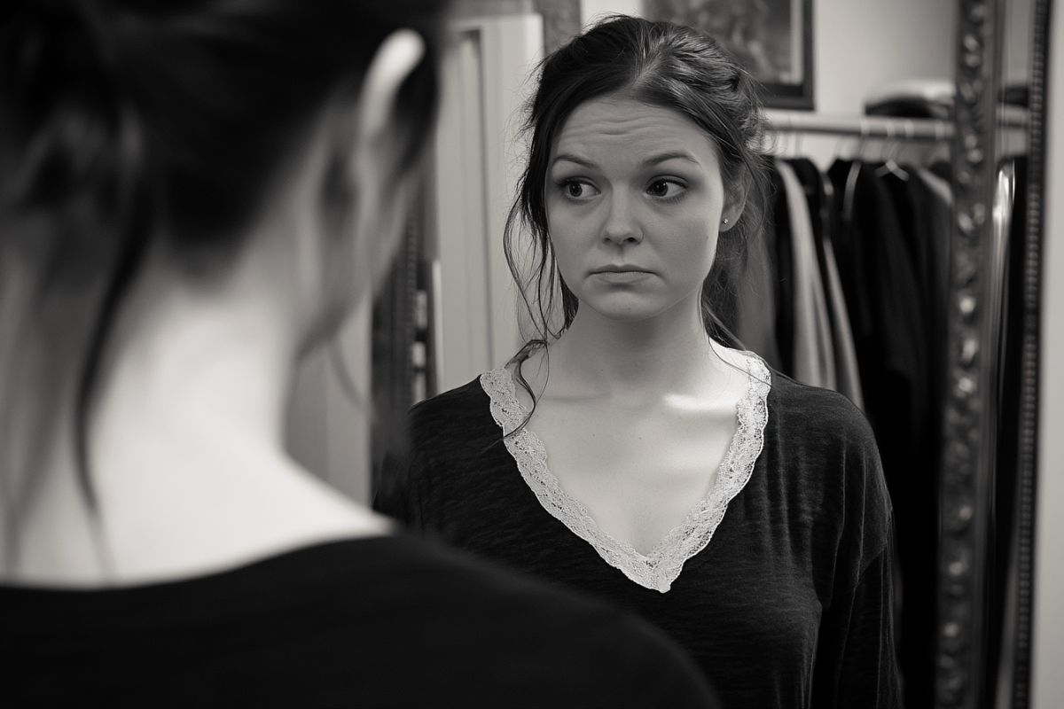 The woman stares in the mirror because she feels sad about the changes in her breasts as she ages.