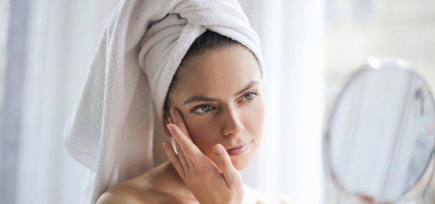 Young woman with a towel on her head touching her face in front of a mirror, showing smooth skin results from BBL halo laser treatment